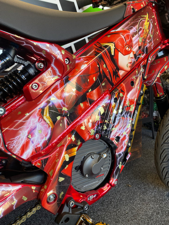 Close-up of a red motorcycle or scooter body panel featuring a detailed Flash comic-style graphic with lightning effects, shown in a workshop setting.