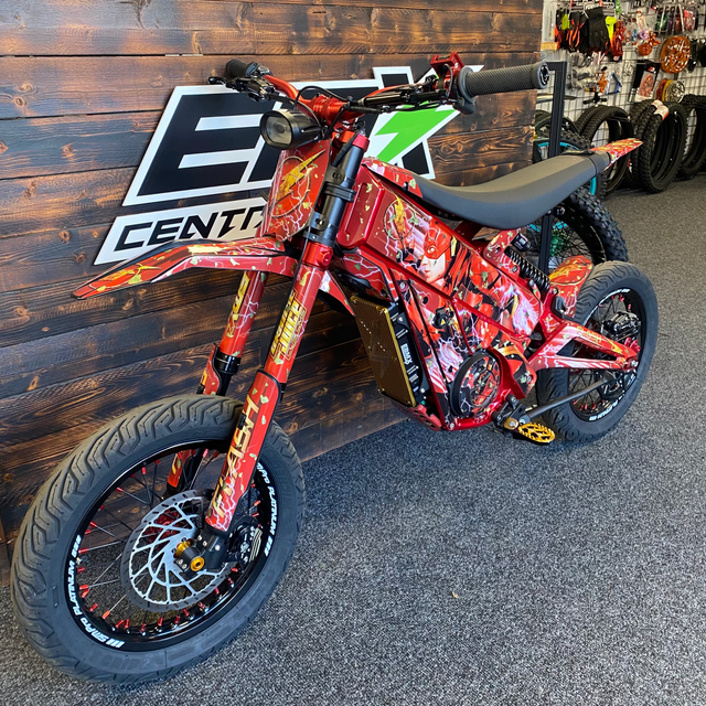 A red electric dirt bike with a colorful flame and graphic wrap, parked indoors against a dark wood wall with a large 'EZ Center' logo.