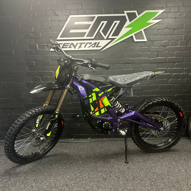 Purple electric bike with colorful design on a stand against a black brick wall with 'EMX' branding.