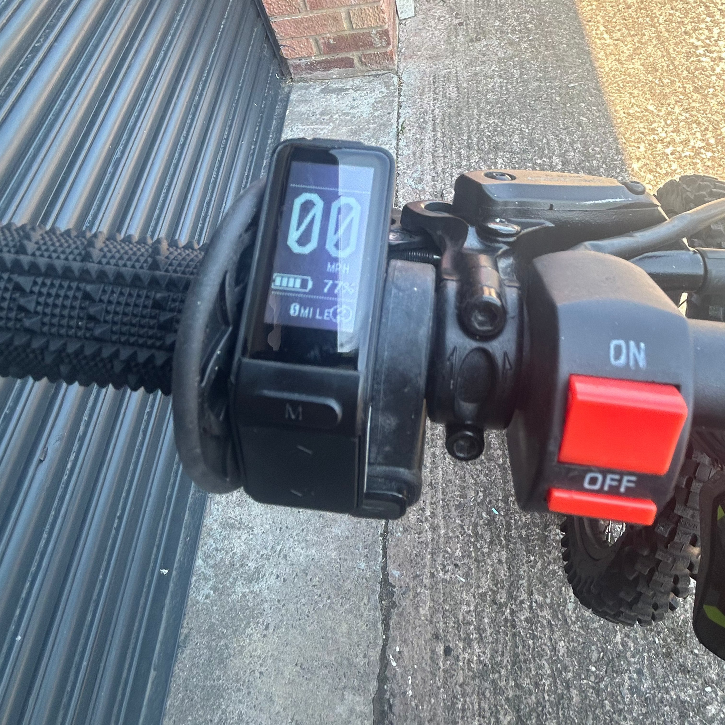 Close-up of a black electric bicycle handlebar with a digital display screen showing '00' and a red power switch labeled 'ON' and 'OFF', set against a real-world outdoor background with a brick wall, metal shutter, and concrete pavement.