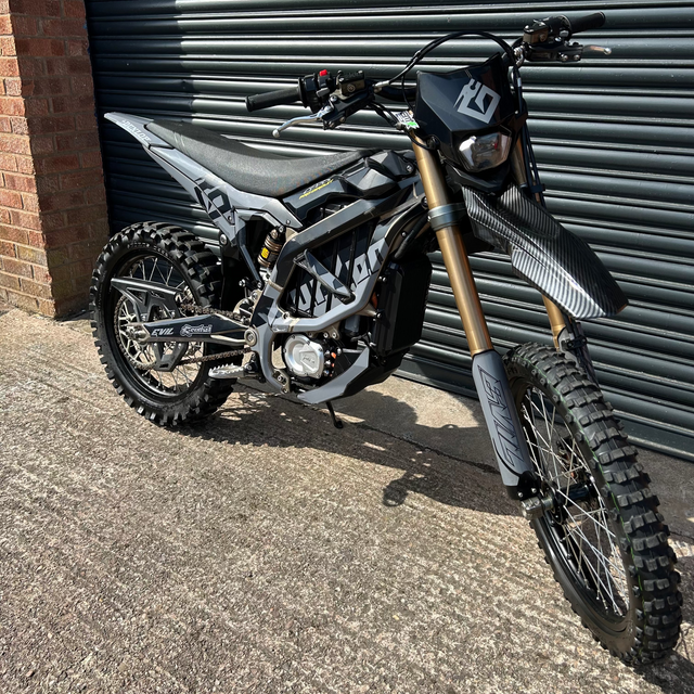 A black and silver electric dirt bike with gold front forks, parked outdoors against a dark gray metal garage door.