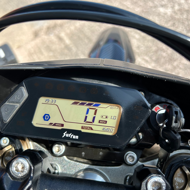 Close-up of a black digital motorcycle instrument cluster with a beige display screen, mounted on a handlebar. The brand name 'Sufan' is visible below the screen.