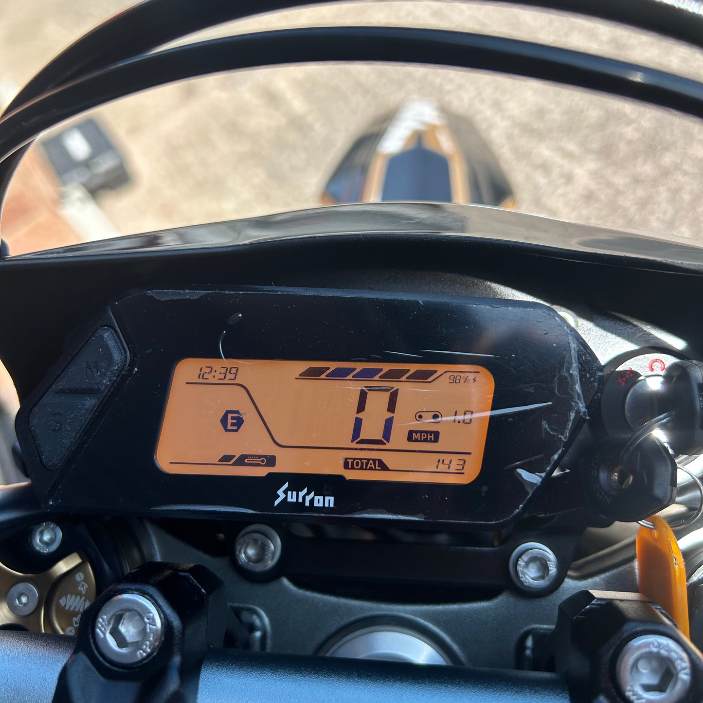 Close-up of a motorcycle dashboard display showing the odometer and instrument panel, mounted on the handlebars with a blurred outdoor background.