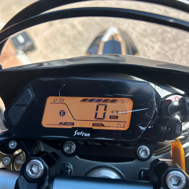 Close-up of a motorcycle dashboard display showing the odometer and instrument panel, mounted on the handlebars with a blurred outdoor background.