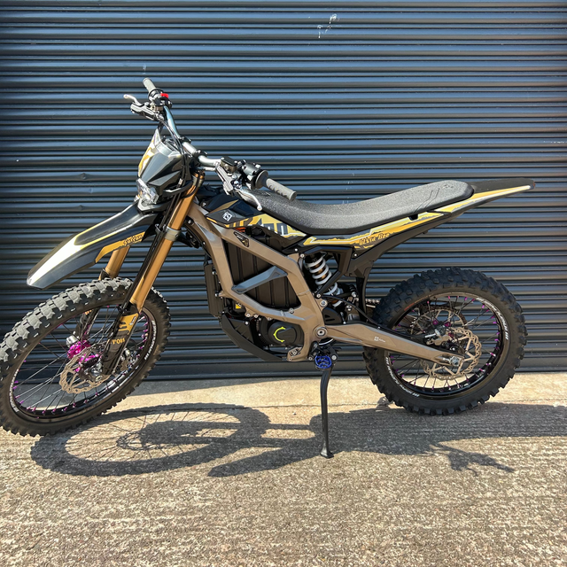 A dirt bike with a black and gold frame, black seat, and knobby tires, parked on a concrete ground in front of a dark gray metal garage door.