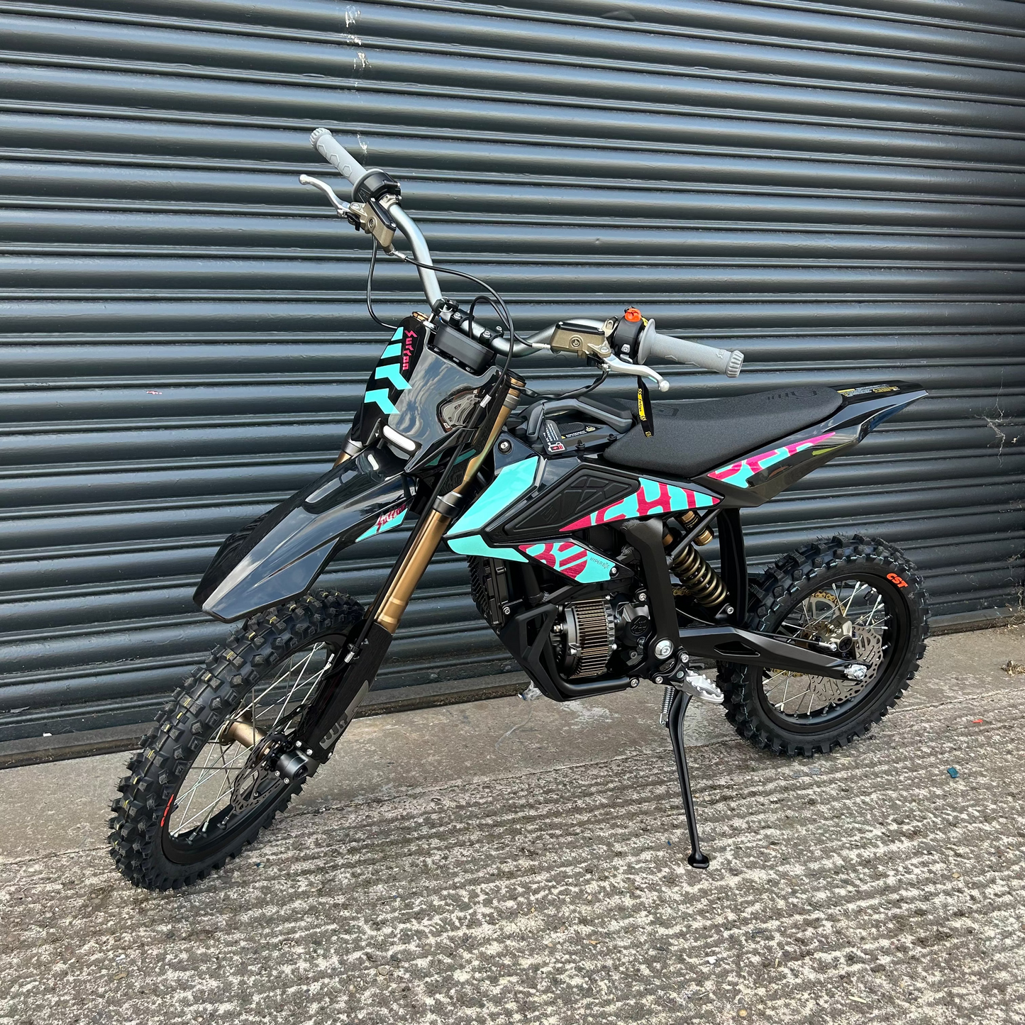 A small off-road dirt bike with black and gray bodywork, gold front fork, and turquoise and pink graphic decals, parked on concrete in front of a gray corrugated metal shutter.