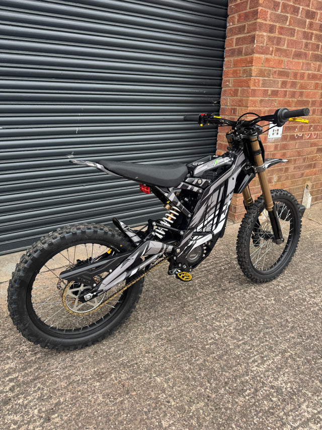 A black and silver electric dirt bike with a gold front fork, parked on concrete next to a brick wall and a dark metal garage door.