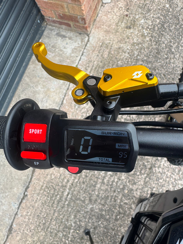 Close-up of a motorcycle or scooter handlebar with a black digital display, red SPORT and EP buttons, and a bright gold brake lever mounted on a black handlebar. The background shows an outdoor concrete surface and brick wall.