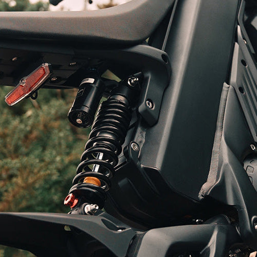 Close-up detail shot of a black motorcycle's rear suspension, featuring a black coil spring with orange and silver accents, a red taillight, and the black frame against a blurred outdoor background.