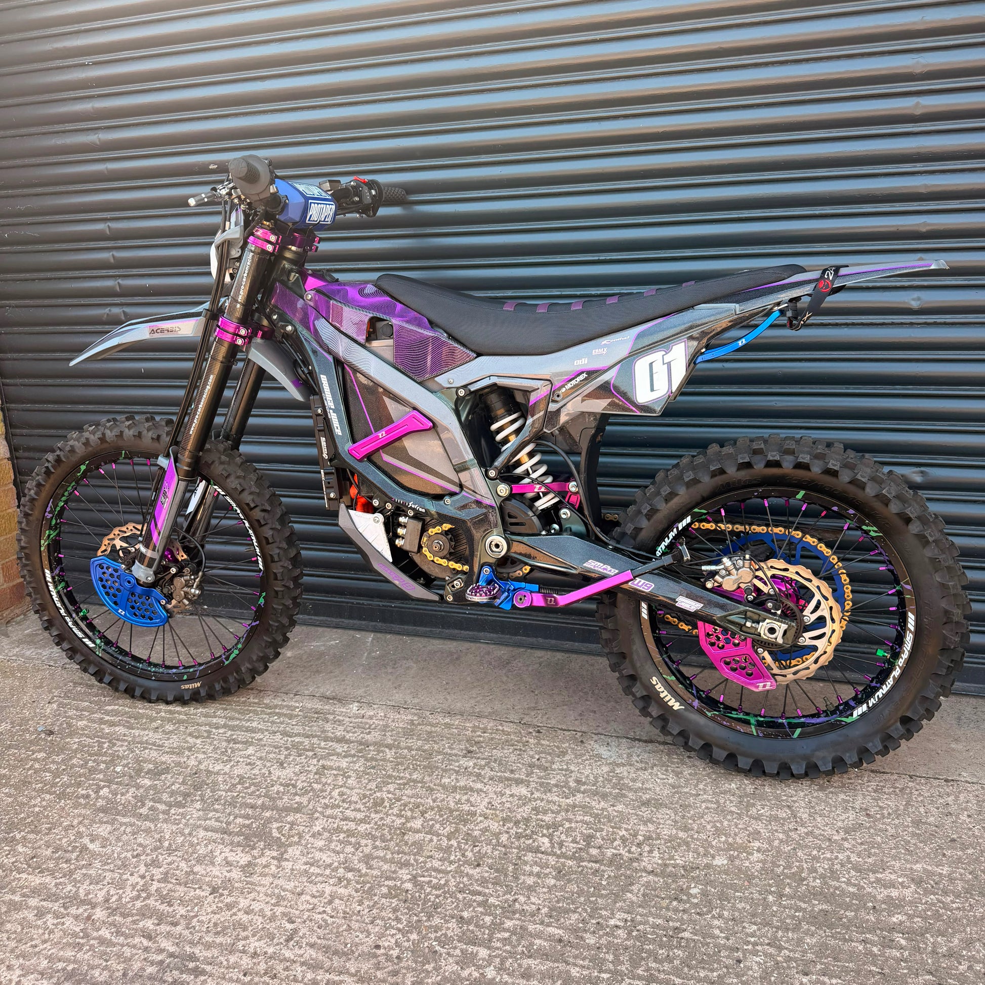 A custom electric dirt bike with a metallic iridescent gray body, purple and pink graphics, and blue and gold wheels, parked in front of a gray metal garage door.