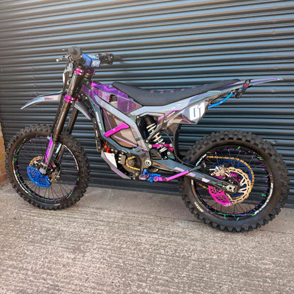 A custom electric dirt bike with a metallic iridescent gray body, purple and pink graphics, and blue and gold wheels, parked in front of a gray metal garage door.