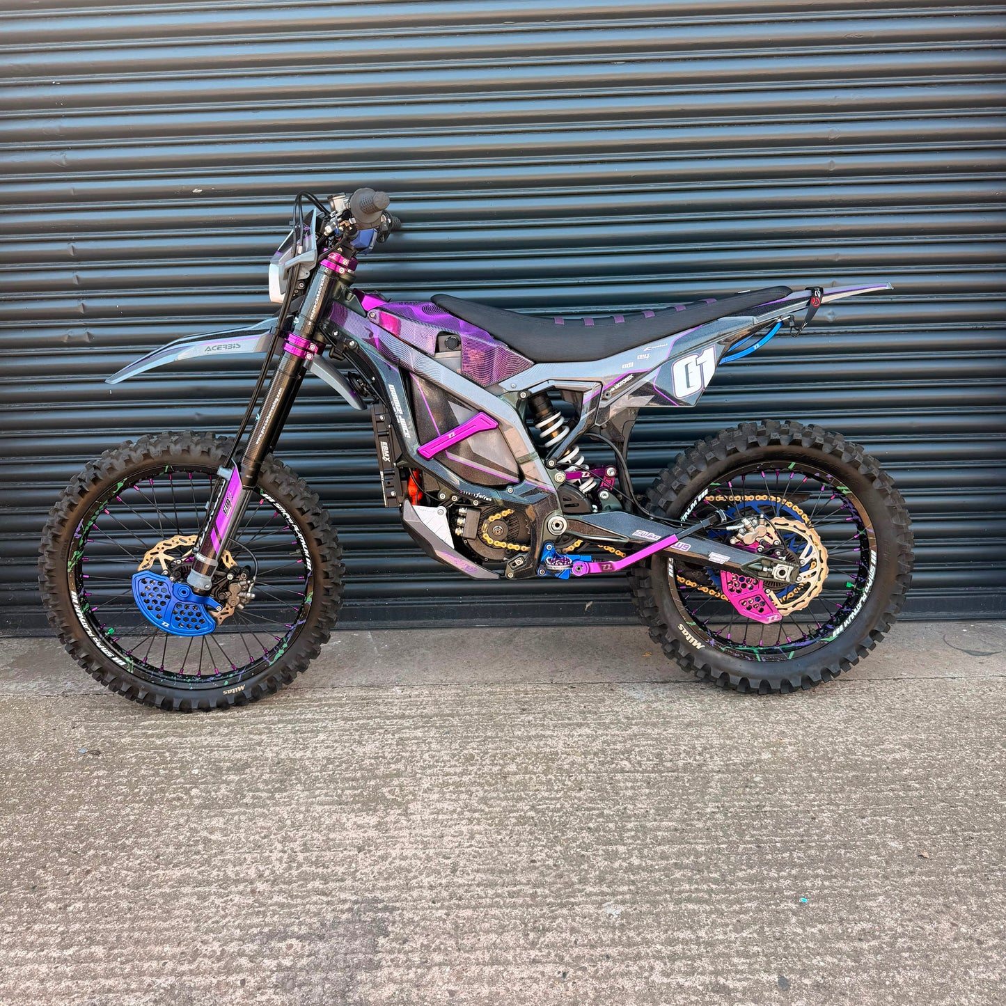 A custom electric dirt bike with a metallic silver and black frame, decorated with vibrant purple, pink, and blue graphics, parked against a gray corrugated metal garage door.
