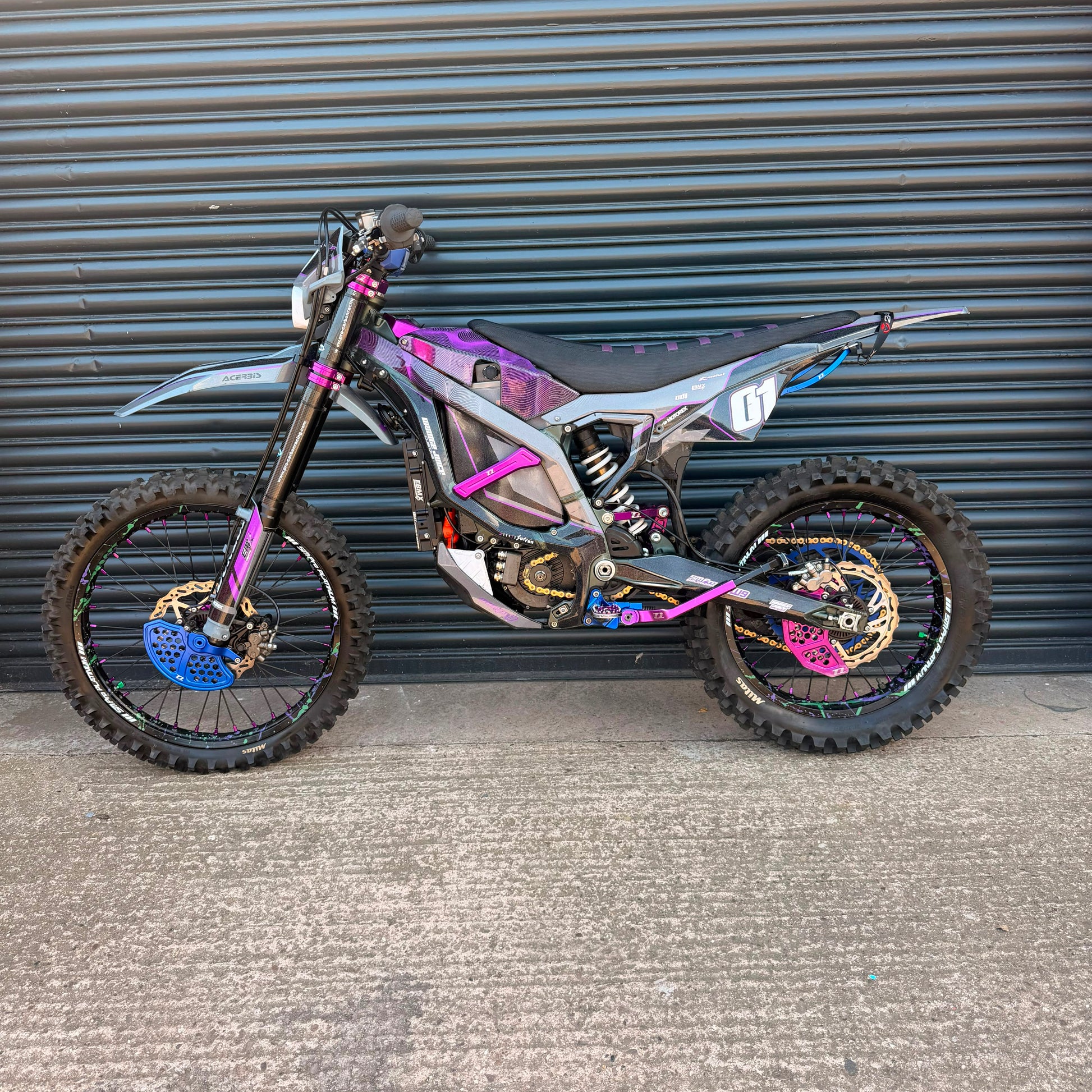 A custom electric dirt bike with a metallic silver and black frame, decorated with vibrant purple, pink, and blue graphics, parked against a gray corrugated metal garage door.