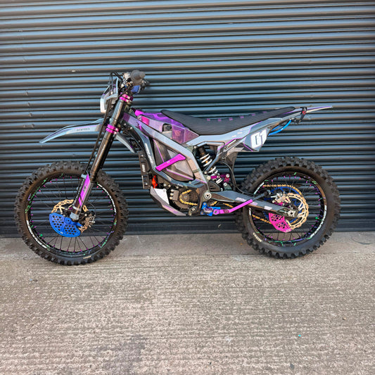 A custom electric dirt bike with a metallic silver and black frame, decorated with vibrant purple, pink, and blue graphics, parked against a gray corrugated metal garage door.