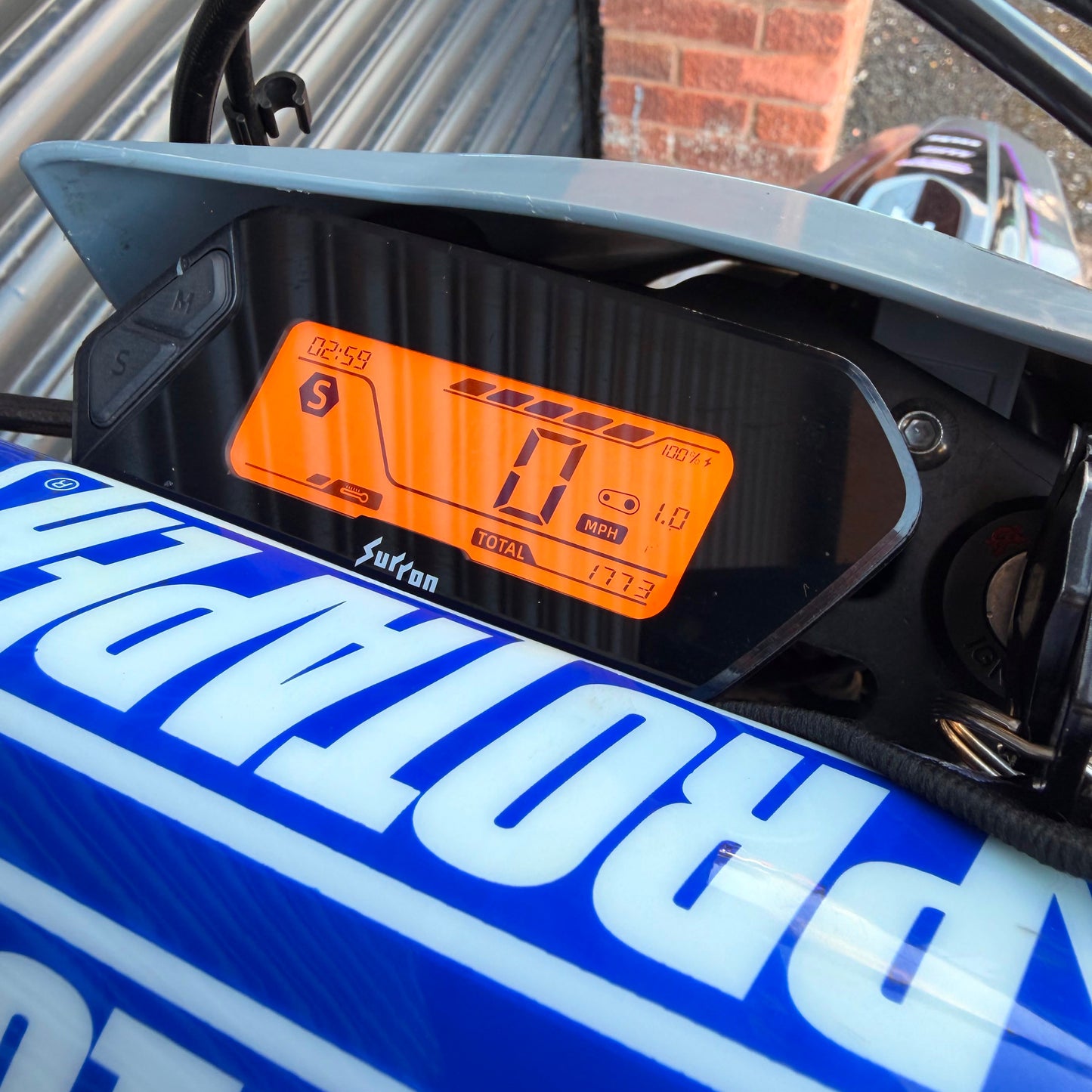 Close-up of a black and gray motorcycle instrument cluster with an orange backlit display, mounted above a blue and white Protaper tank cover.