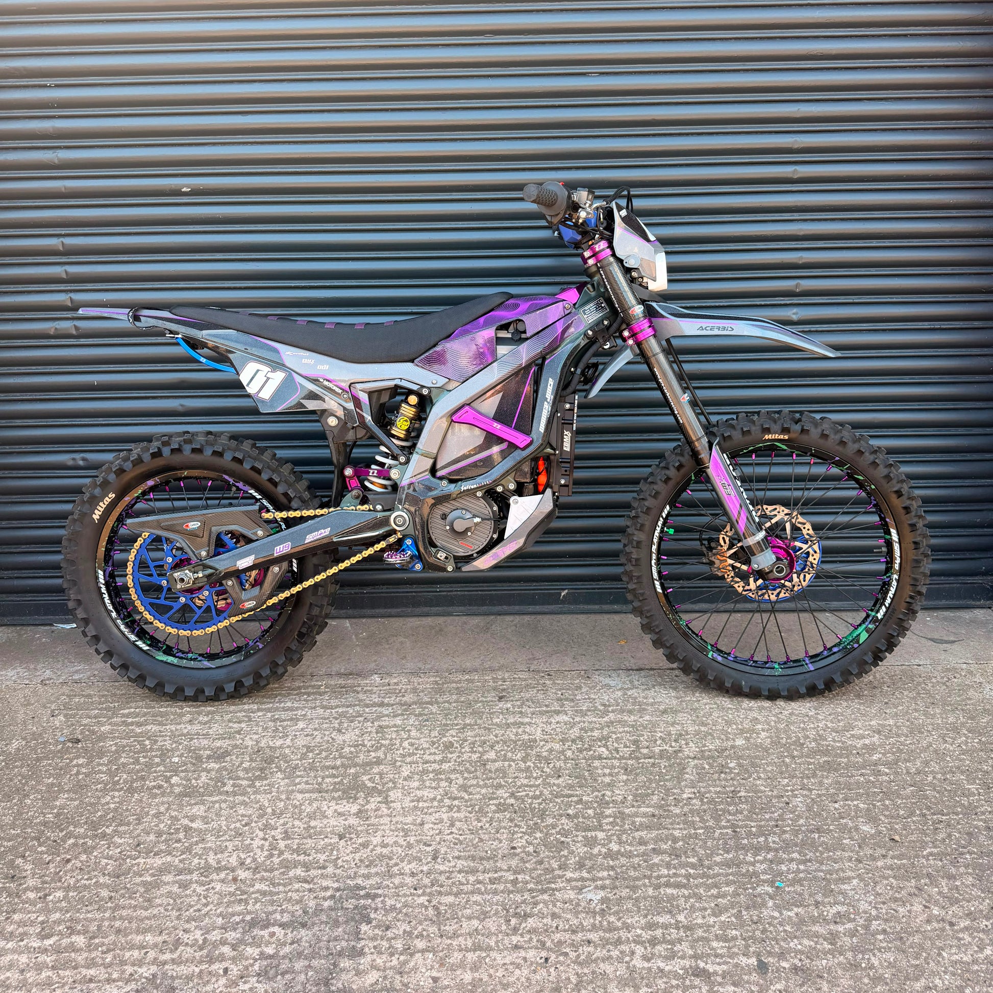 A custom electric dirt bike with a purple and black flame-patterned body, parked against a dark gray corrugated metal garage door.