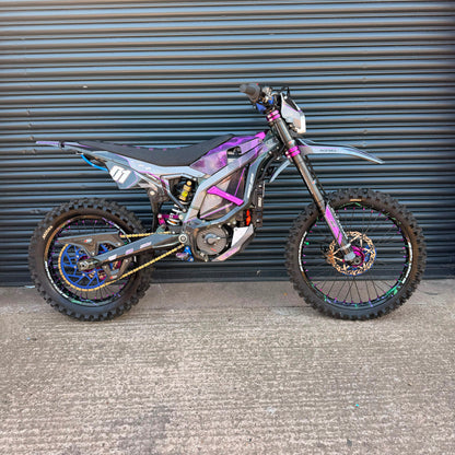 A custom electric dirt bike with a purple and black flame-patterned body, parked against a dark gray corrugated metal garage door.