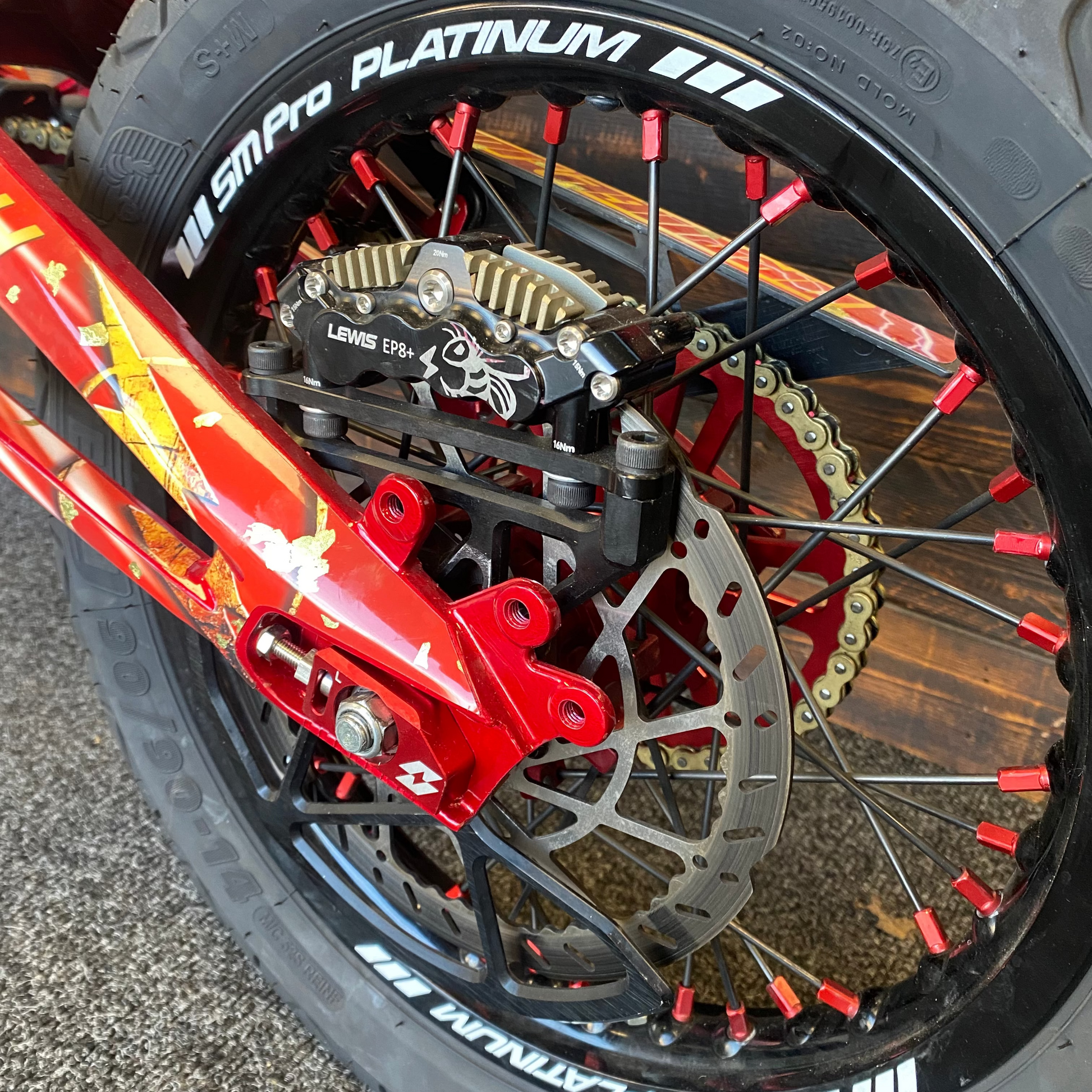 Close-up of a motorcycle wheel featuring a black SMPro Platinum tire, a red brake caliper, a silver disc brake, and red spade-style wheel spacers.