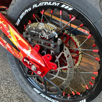 Close-up of a motorcycle wheel featuring a black SMPro Platinum tire, a red brake caliper, a silver disc brake, and red spade-style wheel spacers.