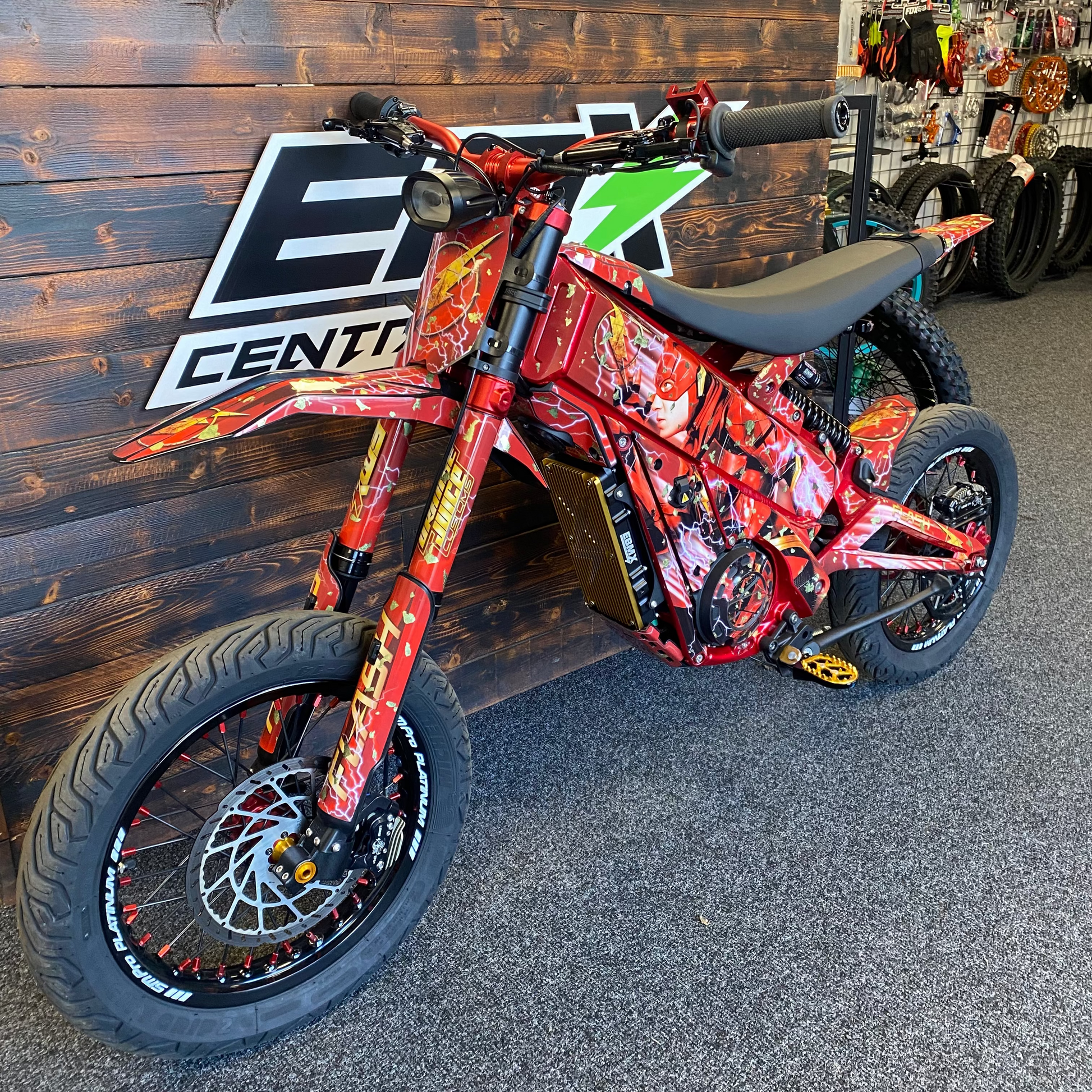 A red electric dirt bike with a colorful flame and graphic wrap, parked indoors against a dark wood wall with a large 'EZ Center' logo.