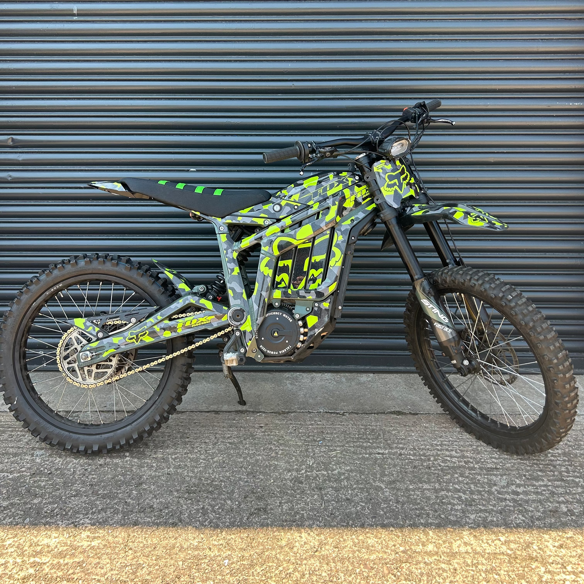 A custom electric dirt bike with a black and neon yellow-green camouflage pattern, parked against a gray metal garage door.