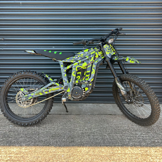 A custom electric dirt bike with a black and neon yellow-green camouflage pattern, parked against a gray metal garage door.