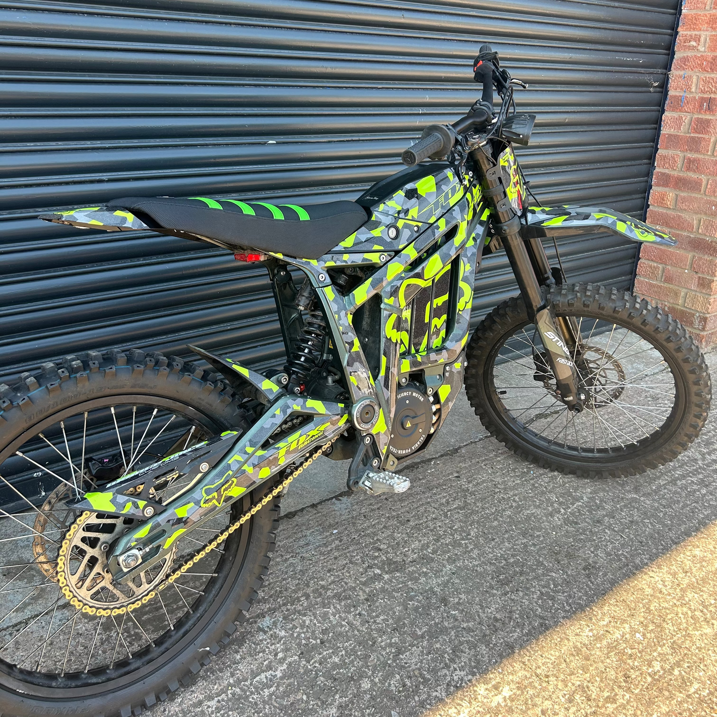 A custom electric dirt bike with a vibrant lime green and black camouflage pattern, parked against a dark gray metal garage door.