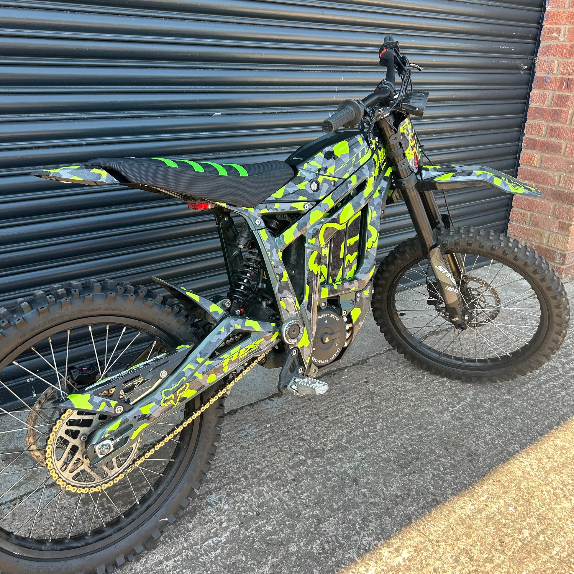 A custom electric dirt bike with a vibrant lime green and black camouflage pattern, parked against a dark gray metal garage door.