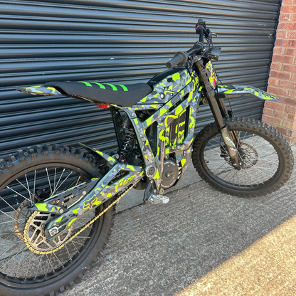 A custom electric dirt bike with a vibrant lime green and black camouflage pattern, parked against a dark gray metal garage door.