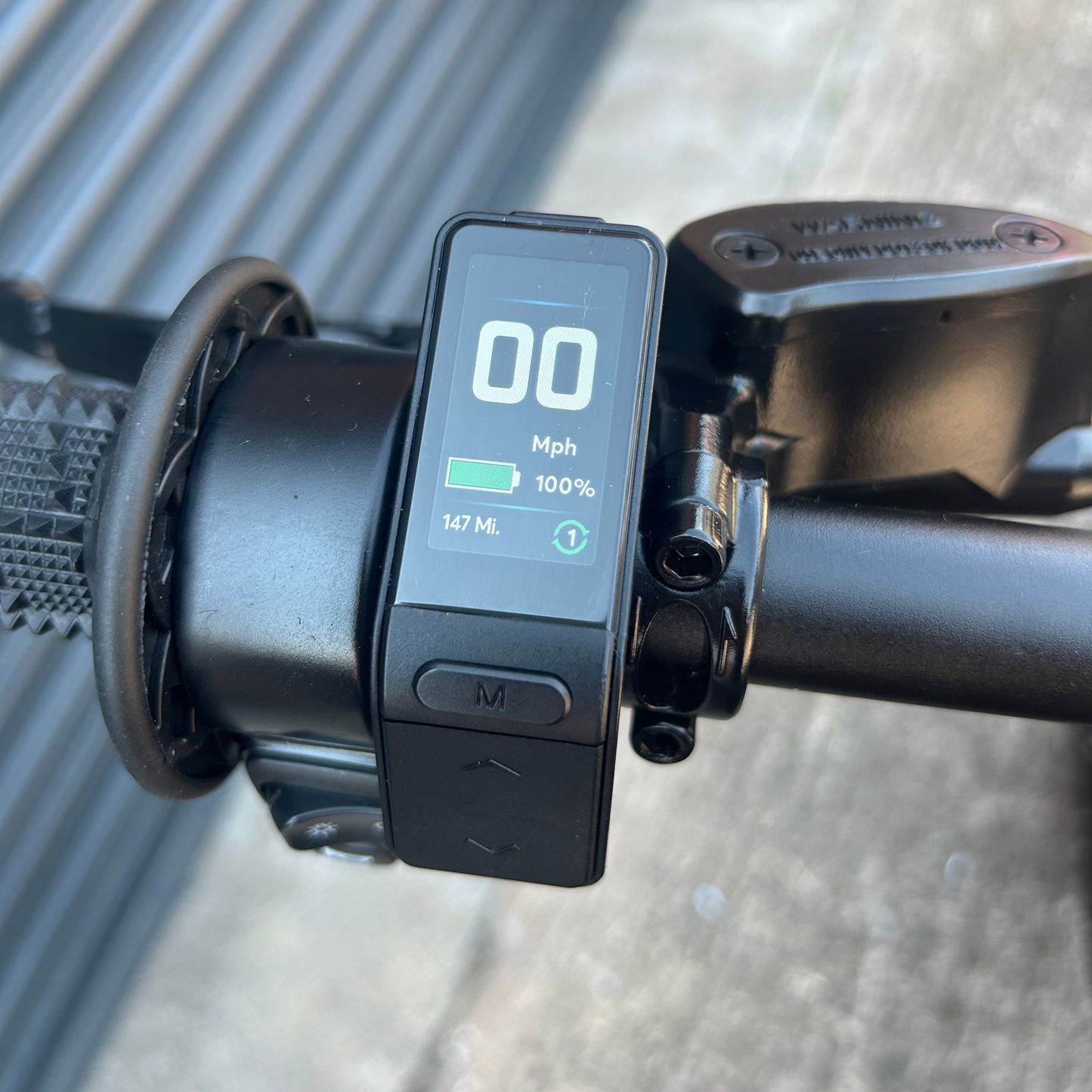 Close-up of a black electric bicycle motor controller with a digital display screen mounted on the handlebar, showing speed and battery percentage, with a blurred outdoor background.