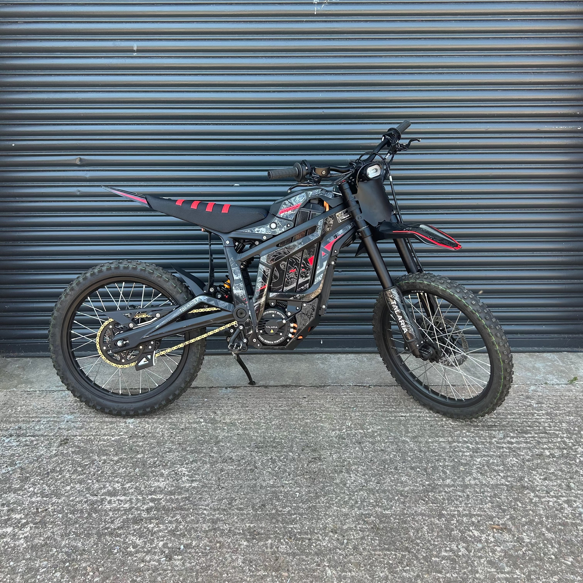 A black and red dirt bike with knobby tires is parked against a dark gray corrugated metal garage door on a concrete ground.