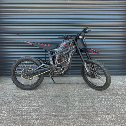A black and red dirt bike with knobby tires is parked against a dark gray corrugated metal garage door on a concrete ground.