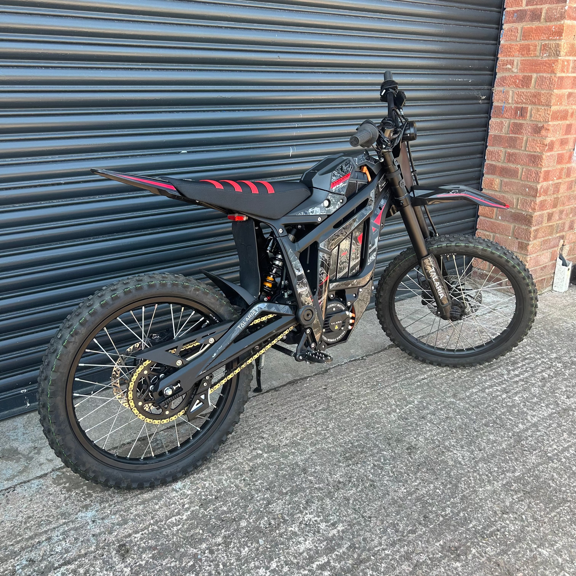A black electric dirt bike with red graphic accents and gold chain drive, parked against a gray metal garage door next to a brick wall.