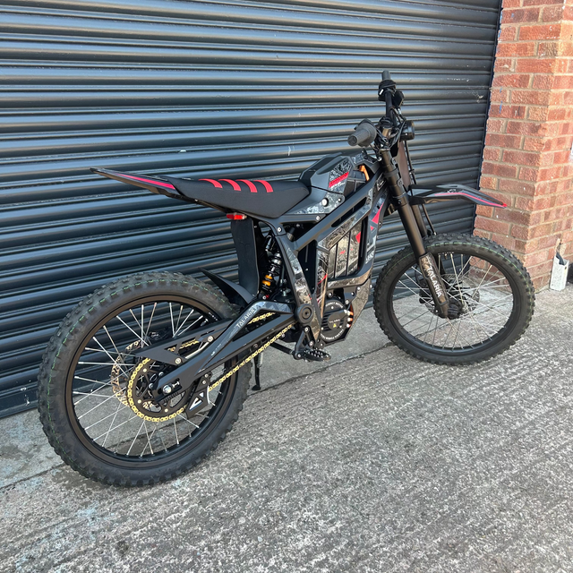 A black electric dirt bike with red graphic accents and gold chain drive, parked against a gray metal garage door next to a brick wall.