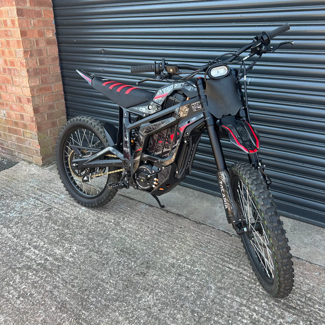 A custom-built electric dirt bike with a black frame, knobby tires, and red and black seat details, parked on concrete in front of a dark gray metal garage door next to a brick wall.
