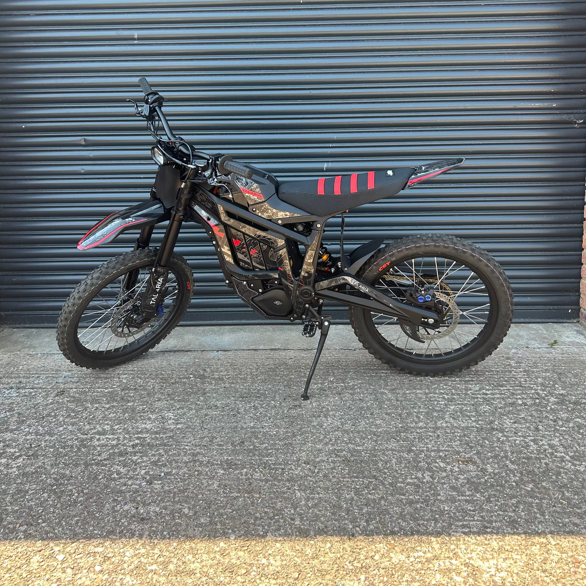 A black electric dirt bike with red and white graphic accents, parked on its kickstand in front of a dark gray corrugated metal garage door.