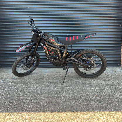 A black electric dirt bike with red and white graphic accents, parked on its kickstand in front of a dark gray corrugated metal garage door.