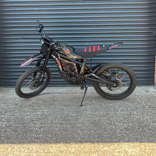 A black electric dirt bike with red and white graphic accents, parked on its kickstand in front of a dark gray corrugated metal garage door.