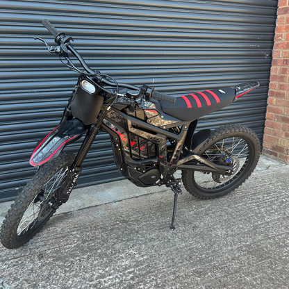 A black dirt bike with red accents on the seat and front fender, parked on a concrete surface in front of a gray metal garage door and a brick wall.