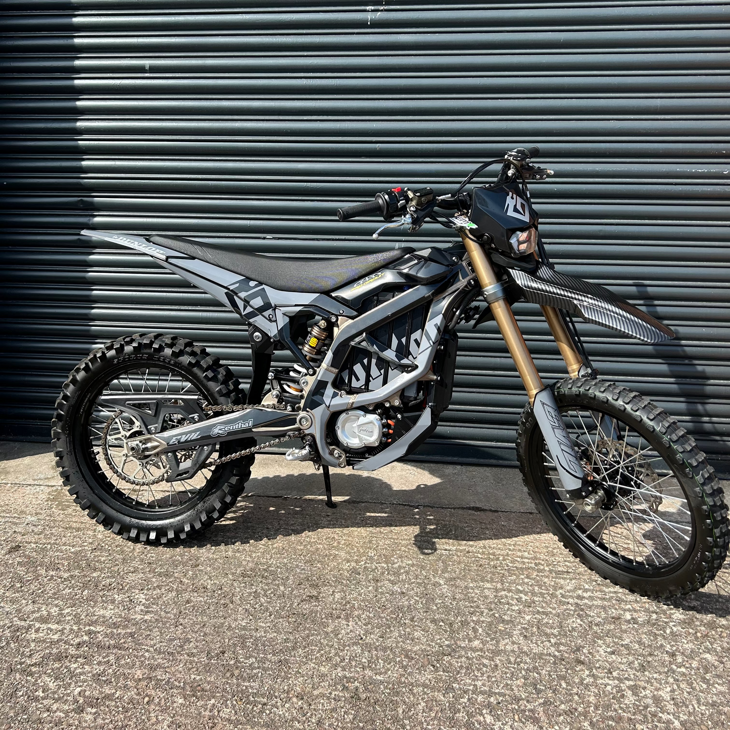 A black and silver dirt bike with gold front forks parked on concrete in front of a dark gray corrugated metal wall.