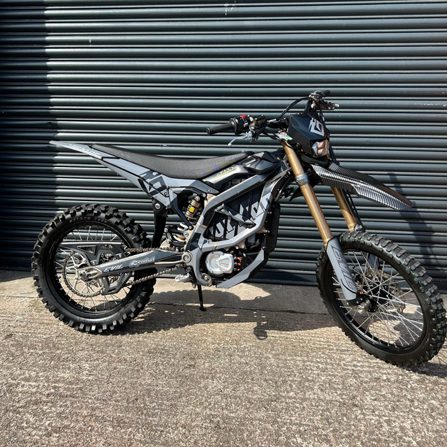 A black and silver dirt bike with gold front forks parked on concrete in front of a dark gray corrugated metal wall.