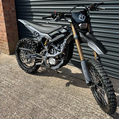 A black and silver electric dirt bike with gold front forks, parked outdoors against a dark gray metal garage door.