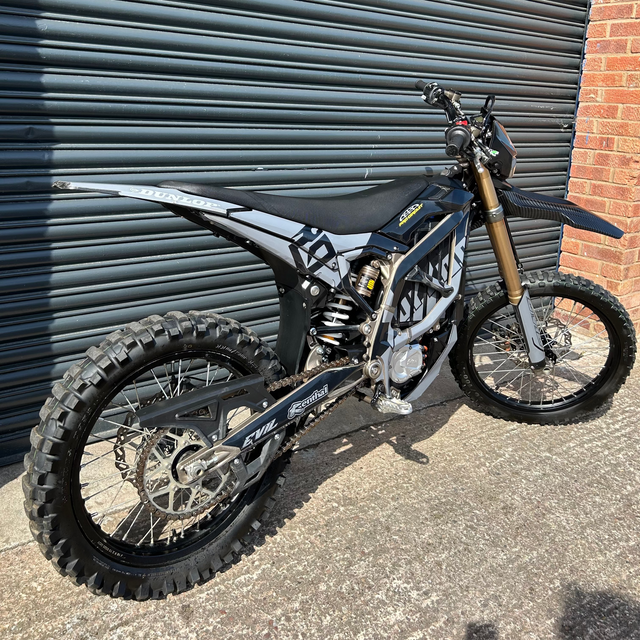A black and silver Husqvarna dirt bike with a gold front fork, parked on a concrete surface in front of a dark gray metal garage door and a red brick wall.