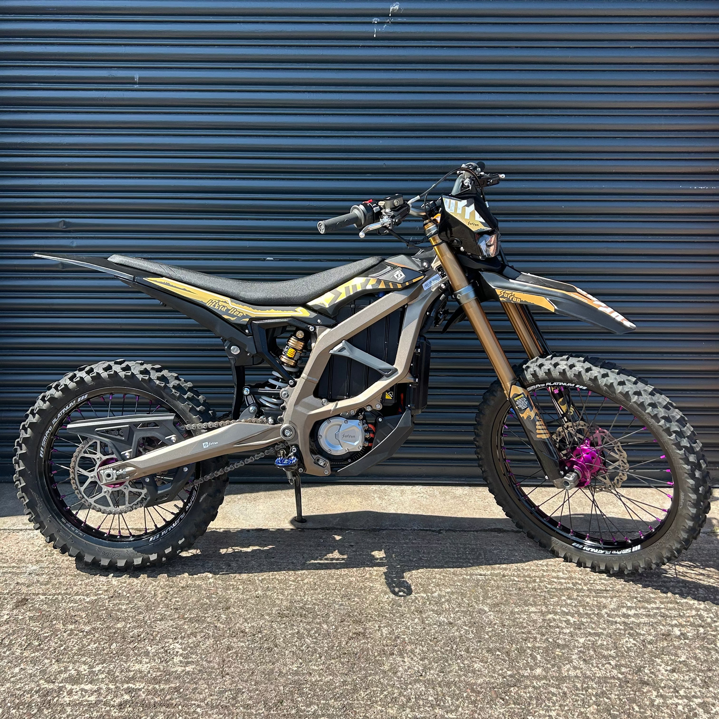 An electric dirt bike with a black and gold frame, gold front fork, and knobby tires, parked on asphalt in front of a dark blue corrugated metal garage door.