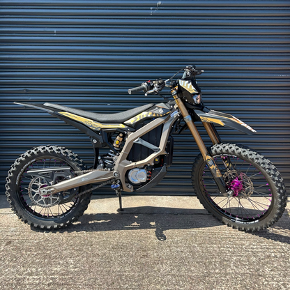 An electric dirt bike with a black and gold frame, gold front fork, and knobby tires, parked on asphalt in front of a dark blue corrugated metal garage door.