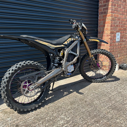 A black and gold dirt bike with knobby off-road tires parked on concrete in front of a dark metal garage door and a red brick wall.