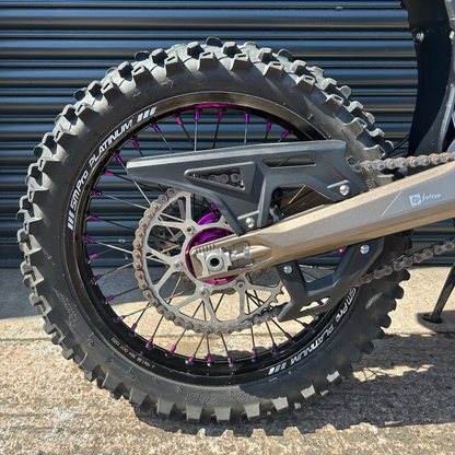 Close-up of a rugged off-road motorcycle rear wheel with a knobby tire, disc brake, chain guard, and purple wheel spokes in front of a dark metal garage door.