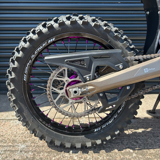 Close-up of a rugged off-road motorcycle rear wheel with a knobby tire, disc brake, chain guard, and purple wheel spokes in front of a dark metal garage door.