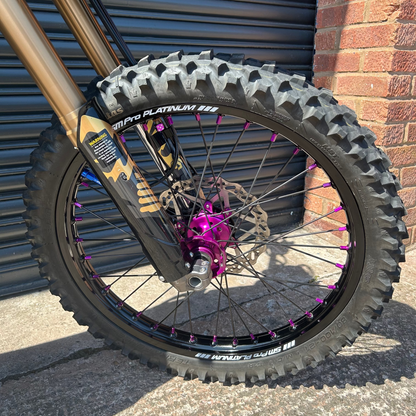 Close-up of a mountain bike front wheel with a knobby black tire, black rim, bright pink wheel spindle, and gold front fork, parked outdoors against a gray metal shutter and brick wall.