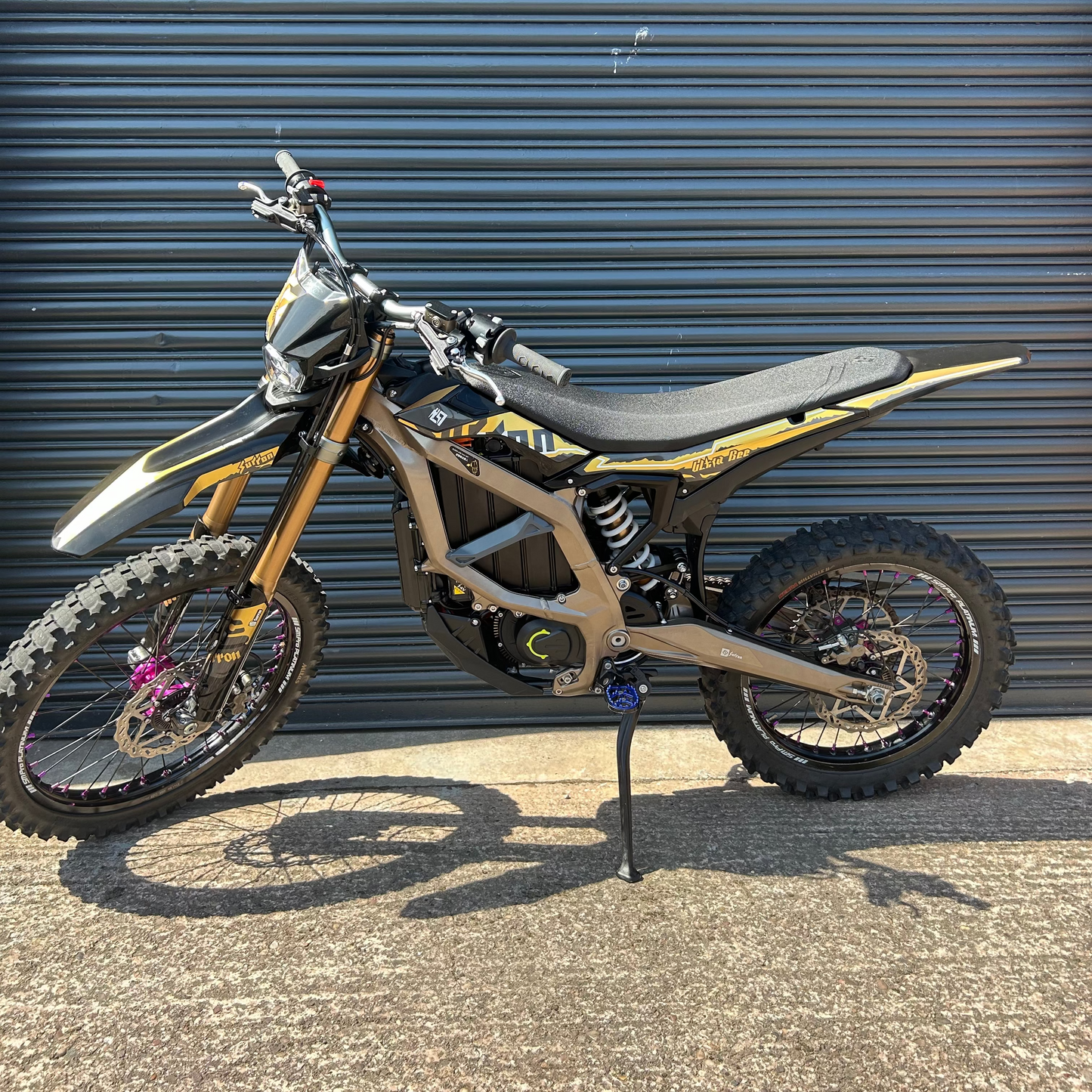 A dirt bike with a black and gold frame, black seat, and knobby tires, parked on a concrete ground in front of a dark gray metal garage door.